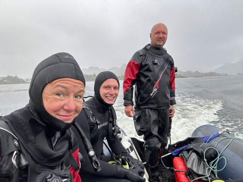 Group of three scuba divers, one woman, two men, sitting in a boat suited up for cold water diving.