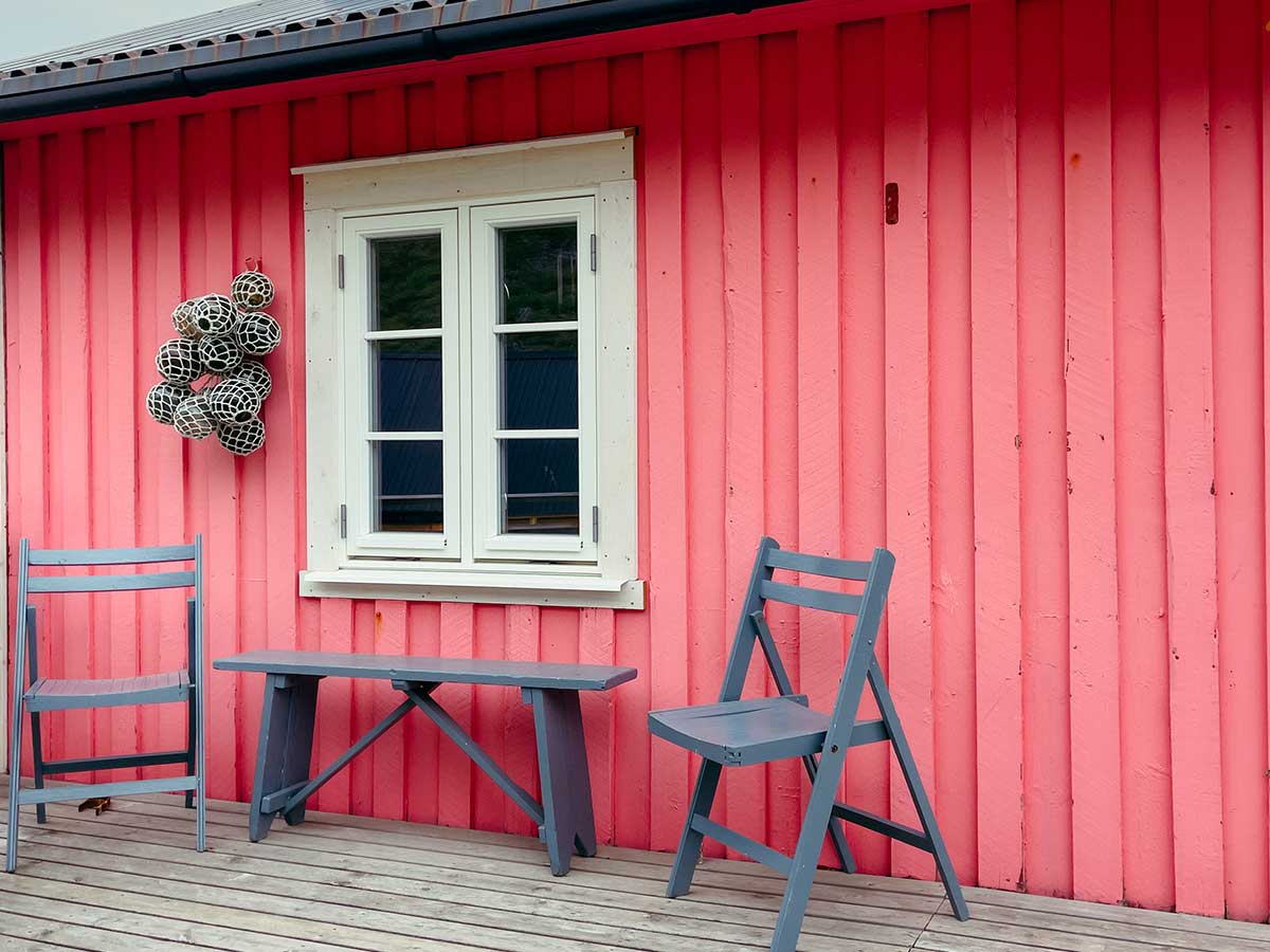Red wooden cabin facade with white window and two grey chairs on the deck.