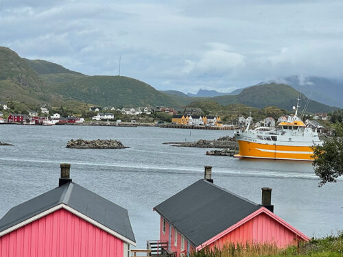 View of Ballstad from Havly Lofoten retreat, showing two pink cabins in front of the picture. A big yellow and white ferry enters the picture from the right.